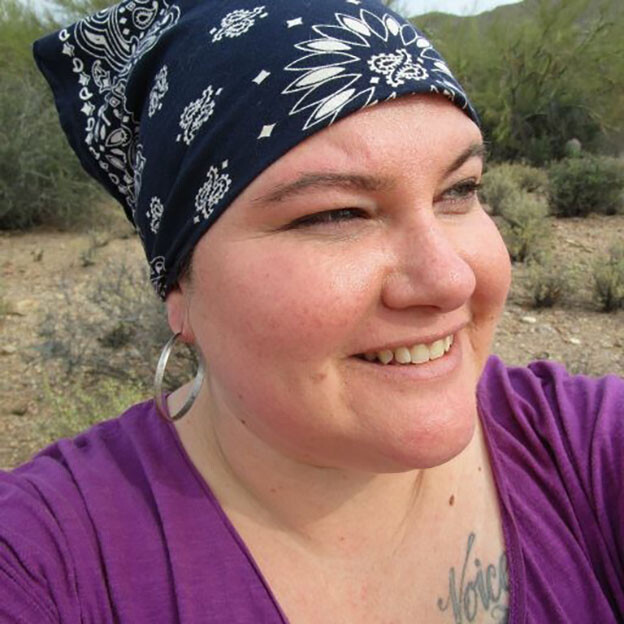 A white Mestiza woman stands in the desert wearing a purple top and blue bandana